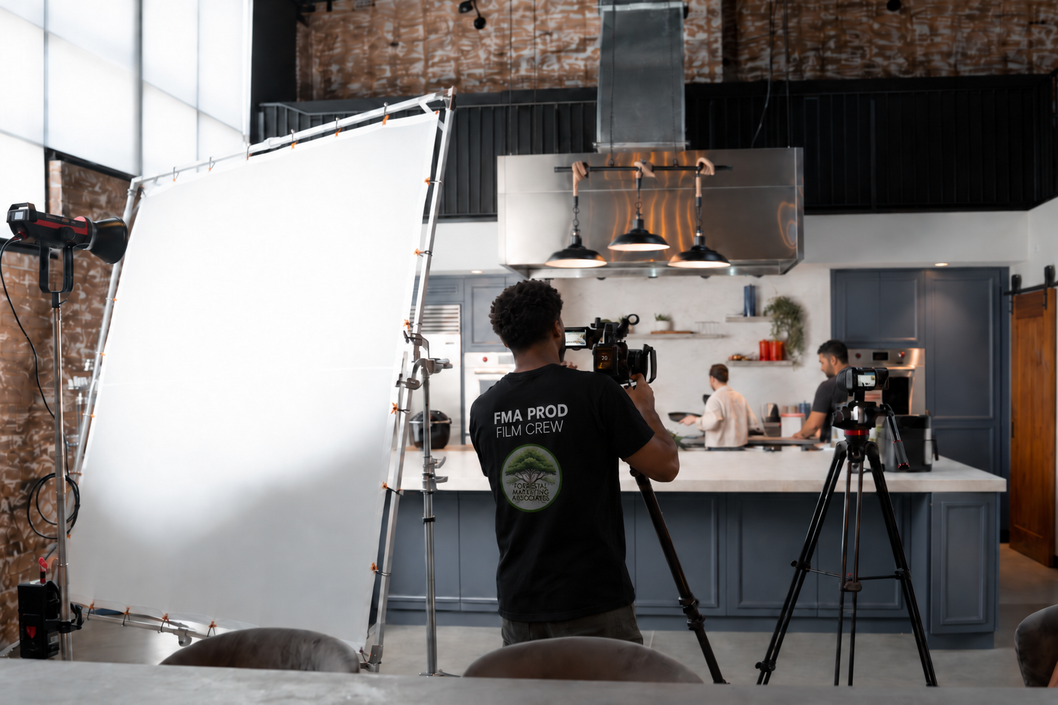 An FMA film crew member operating a camera on a kitchen production set, with lighting equipment and a second camera